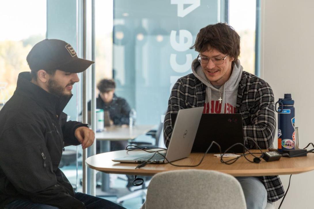 Two students sit at a small table looking at laptops